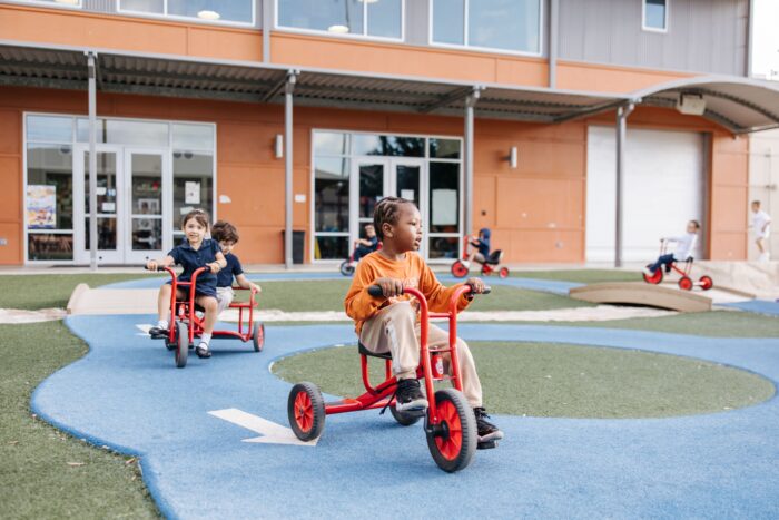 A student on a single trike and two students on a double trike riding on the playground